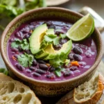 A bowl of black bean soup garnished with avocado slices, cilantro, and a lime wedge, with slices of bread on the side.